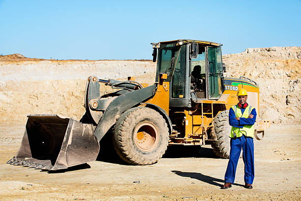 Full length portrait of quarry worker standing arms crossed by bulldozer at construction site